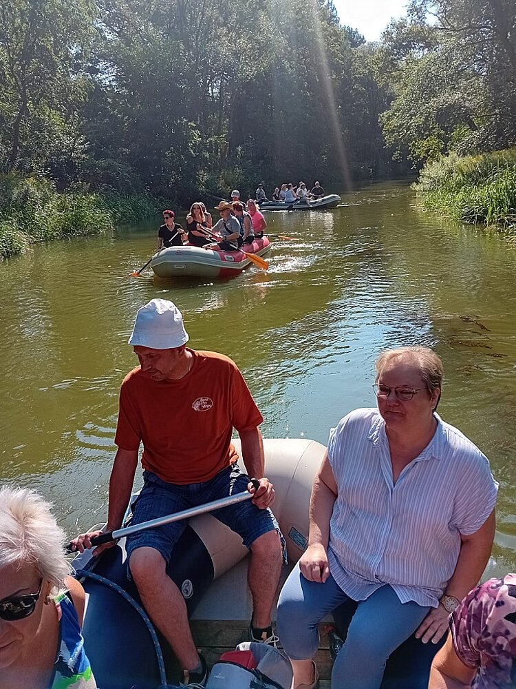 Drei Boote fahren auf einem kleinen Fluss. Das Bild ist vom ersten Schlauchboot aus aufgenommen, darin sieht man drei Personen sitzen. Das zweite Boot fährt in Fließrichtung des Stroms, das dritte hingegen steht quer zum Fluss.