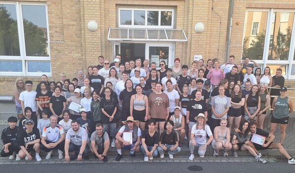 Ein große Gruppe von Schülerinnen, Schülern und Lehrkräften in Sportkleidung hat sich zu einem Gruppenbild vor dem Eingang des Schulgebäudes versammelt.