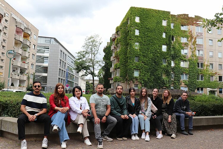 Unser Integrationskurs besucht Leipzig Eine Gruppe von zehn Personen sitzt auf einer Steinbank vor einem begrünten Wohnhaus in der Stadt.