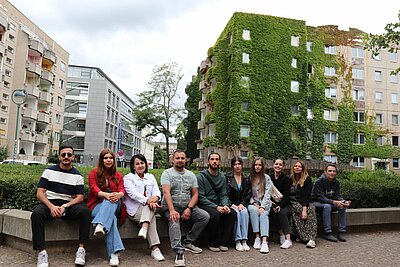 Unser Integrationskurs besucht Leipzig Eine Gruppe von zehn Personen sitzt auf einer Steinbank vor einem begrünten Wohnhaus in der Stadt.