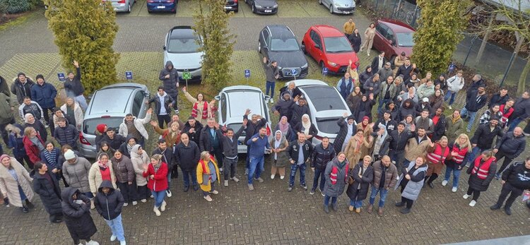 Gruppe von vielen Menschen, insgesamt waren es rund 130 - auf dem Bild sind nicht alle abgebildet, steht während einer Evakuierungsübung auf dem Parkplatz der Euro-Schulen Viersen. Einige tragen rote Warnwesten und schauen winkend nach oben, denn das Bild ist aus der Vogelperspektive aufgenommen. Im Hintergrund sind mehrere geparkte Autos und Bäume zu sehen.