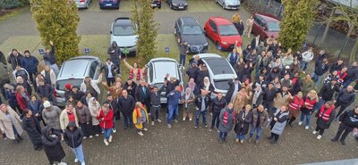 Gruppe von vielen Menschen, insgesamt waren es rund 130 - auf dem Bild sind nicht alle abgebildet, steht während einer Evakuierungsübung auf dem Parkplatz der Euro-Schulen Viersen. Einige tragen rote Warnwesten und schauen winkend nach oben, denn das Bild ist aus der Vogelperspektive aufgenommen. Im Hintergrund sind mehrere geparkte Autos und Bäume zu sehen.
