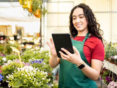 Eine junge Frau mit lockigem Haar und einer grünen Schürze über einem roten Shirt steht in einem Blumenladen. Sie hält ein Tablet in den Händen und lächelt. Um sie herum sind verschiedene Blumen und Pflanzen in Töpfen zu sehen. Der Hintergrund zeigt Regale mit weiteren Blumen und ein helles Gewächshaus.