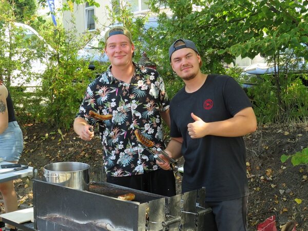 Zwei gute gelaunte, männliche Auszubildende grillen Würstchen für die Besucher*innen.