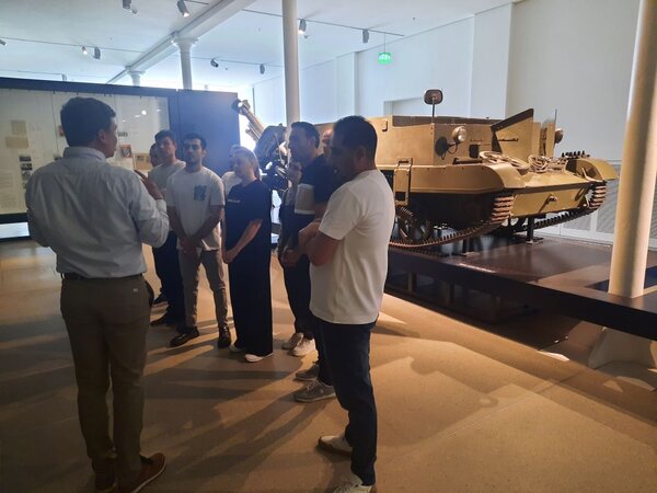 Eine Gruppe von Menschen steht in einem Museum und hört einem Museumsführer zu. Hinter ihnen ist ein Panzer auf einer Plattform ausgestellt.