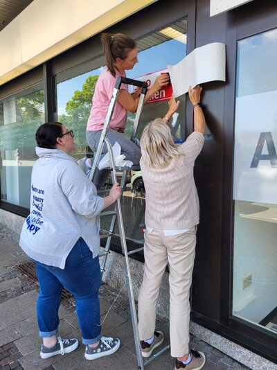 Drei Personen stehen vor einem Gebäude, wovon eine Person auf einer Leiter steht und gemeinsam mit einer anderen einen eine Klebefolie mit der Aufschrift "Euro-Schulen" auf ein Fenster aufkleben. Die dritte Person hält dabei die Leiter fest.