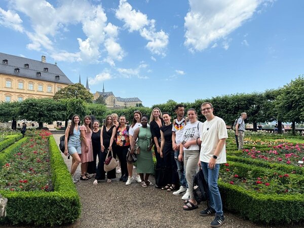 Die Gruppe der Teilnehmenden steht auf einem Kiesweg in einer gepflegten Gartenanlage mit rechteckigen Blumenbeeten, die von niedrigen Buchsbaumhecken eingefasst sind. Im Hintergrund sind dichte, akkurat geschnittene Baumreihen und ein historisches Gebäude mit spitz zulaufenden Türmen zu sehen. Der Himmel ist leicht bewölkt, die Szene wirkt sommerlich und freundlich. Die Personen tragen sommerliche Kleidung und lächeln in Richtung Kamera.