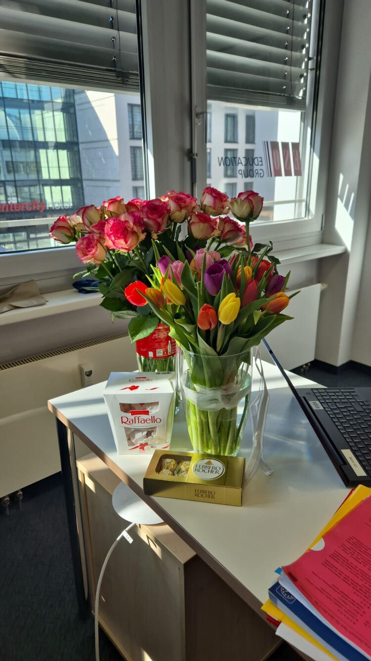 Auf einem Büroschreibtisch stehen neben Laptop und Unterladen zwei Blumensträuße aus Tulpen und Rosen, daneben liegen eine Packung Raffaello und eine Packung Ferrero Rocher.