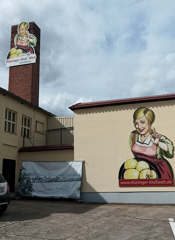 Außenansicht der Thüringer Kloß-Welt in Heichelheim mit großem Wandmotiv einer Frau mit Thüringer Klößen und entsprechendem Schriftzug.