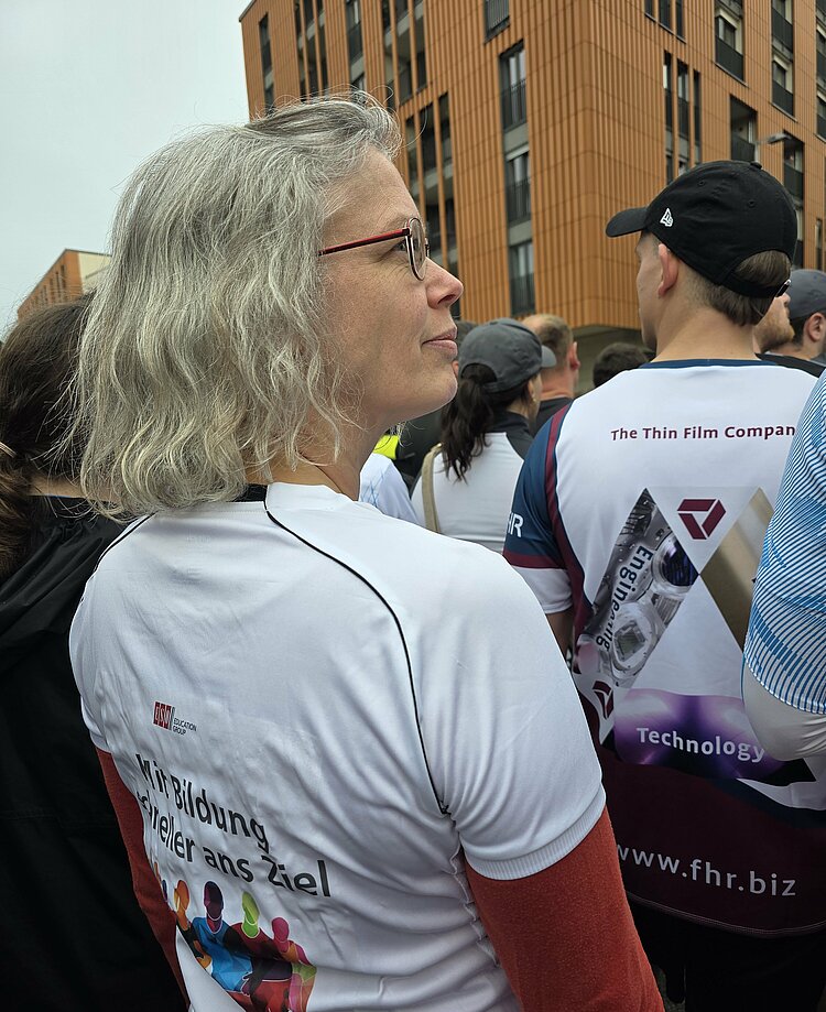 Euro-Schulen & Euro Akademie bei der Rewe Team Challenge 2025 Nahaufnahme einer Frau mit grauen Haaren in Laufkleidung von hinten. Auf dem Shirt steht „Mit Bildung schneller ans Ziel“. Um sie herum sind weitere Läufer zu sehen.