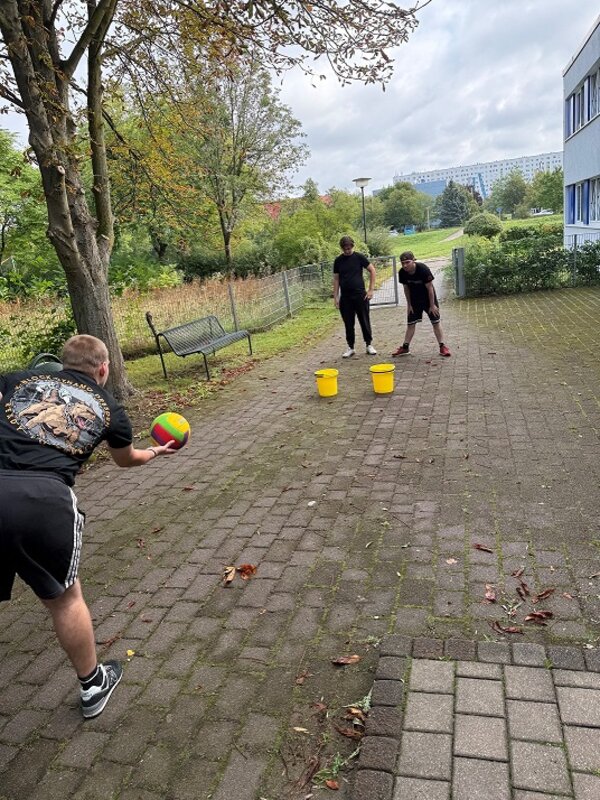 Drei Personen spielen im Freien mit einem Ball und zwei Eimern auf einem gepflasterten Weg.