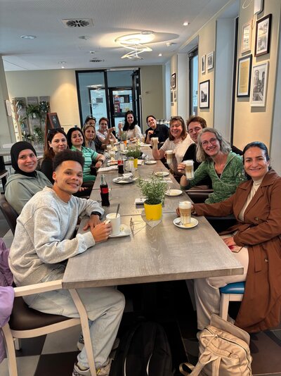 Eine größere Gruppe von Erwachsenen sitzt gemeinsam an einem langen Tisch in einem Café, lächelt in die Kamera und hält teilweise Kaffee- und Latte-Macchiato-Gläser in den Händen.