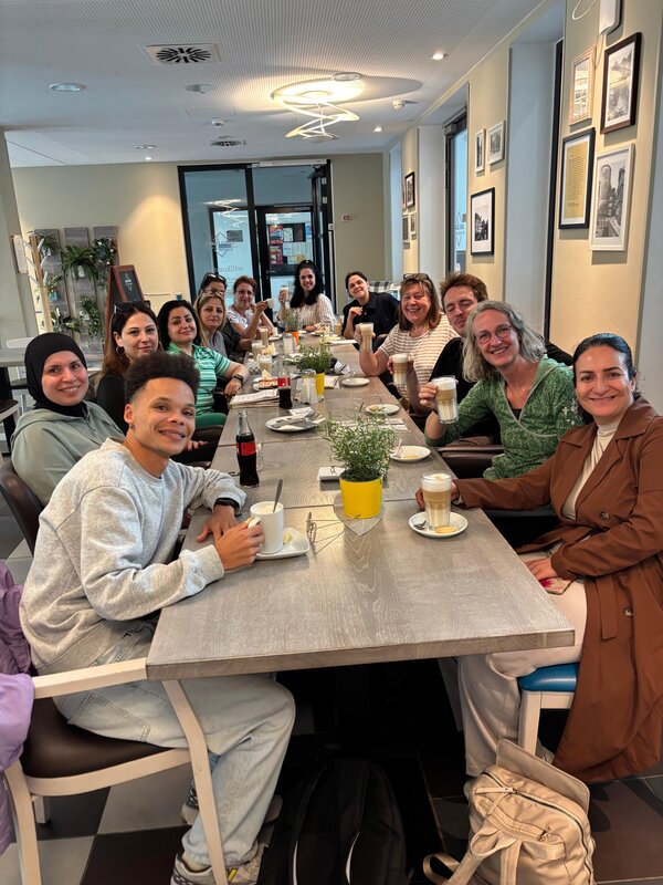Eine größere Gruppe von Erwachsenen sitzt gemeinsam an einem langen Tisch in einem Café, lächelt in die Kamera und hält teilweise Kaffee- und Latte-Macchiato-Gläser in den Händen.