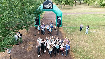 Gemeinsam unterwegs für die Gesundheit Aus der Vogelperspektive fotografiertes Gruppenbild der Mitarbeitenden der Euro-Schulen und Euro Akademie Magdeburg. Sie schauen gut gelaunt und winkend nach oben, stehen auf einer großen Wiese am Start der Firmenwanderung, der als großer, aufgeblasener grüner Bogen aufgestellt ist.