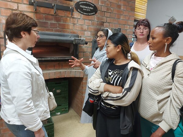 Eine Frau erklärt einer kleinen Gruppe von Frauen die Funktion eines traditionellen Backofens. Die Gruppe steht vor einer Ziegelwand mit Backofenöffnung.