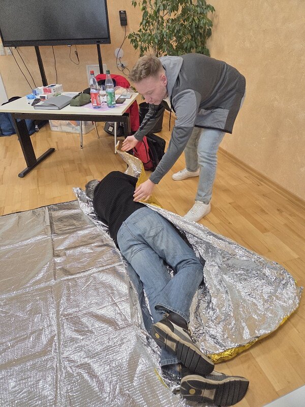 Ein Mitarbeiter der Erste-Hilfe-Schulung deckt den in stabiler Seitenlage liegenden Mitarbeiter mit einer Rettungsdecke zu, um Unterkühlung zu vermeiden. Praktische Übung im Rahmen einer Fortbildung zur Ersten Hilfe.