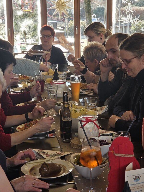 Mehrere Personen sitzen dicht an einem Tisch und essen gemeinsam, auf dem Tisch stehen Getränke, Teller mit warmen Speisen und weihnachtliche Dekoration