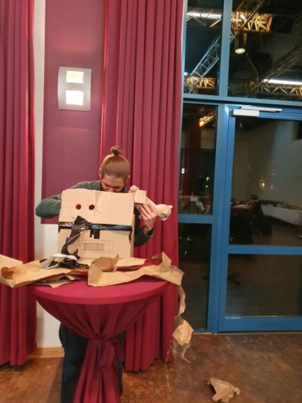 Eine Person steht an einem mit roter Tischdecke bespannten Stehtisch vor einem roten Vorhang und hält ein großes, teilweise ausgepacktes Pappkarton-Objekt in den Händen. Um den Tisch herum liegen Reste von Packpapier auf dem Boden.