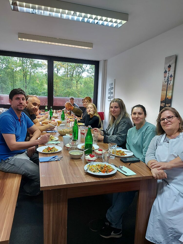 Die Teilnehmenden sitzen gemeinsam an einem großen Tisch vor eine breiten Fenster und sind kurz davor, mit dem Essen zu beginnen. Vor ihnen stehen große Teile mit bunten Zutaten, auch Desserts und Wasserflaschen und -gläser sind auf dem Tisch erkennbar.