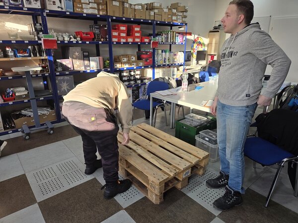 Zwei junge Männer in einem Lagerraum mit Warenregalen und Tischen; einer beugt sich über eine Holzpalette, der andere steht daneben und beobachtet.