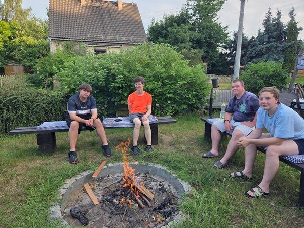 Die vier männlichen Auszubildenden zu Fachlageristen sitzen gemeinsam vor einem kleinen Lagerfeuer, sind sichtlich erschöpft, aber guter Dinge. 