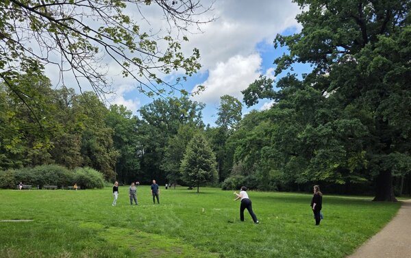 Eine Gruppe von Menschen steht auf einer Wiese in einem Park und führt eine Gruppenaktivität durch. Im Hintergrund sind Bäume und Parkbänke zu sehen. Der Himmel ist leicht bewölkt und die Atmosphäre wirkt entspannt.