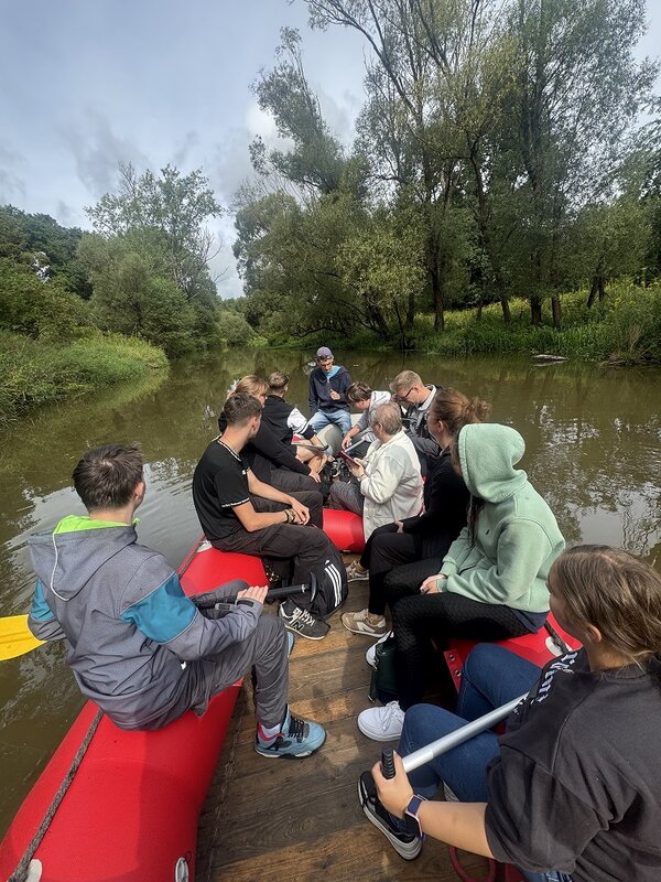 Eine größere Gruppe von Menschen sitzt in einem roten Schlauchboot auf einem ruhigen Fluss. Einige unterhalten sich, während andere in die Ferne schauen. Die Umgebung ist von Bäumen und Sträuchern geprägt.