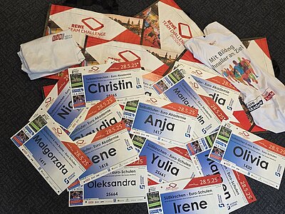 Unsere Teams laufen los – REWE Team Challenge 2025 Startnummern für die REWE Team Challenge mit Namen der Mitarbeitenden der Euro Akademien und Euro-Schulen liegen auf einem Haufen, dazwischen bedruckte Euro Akademie und Euro-Schulen Teilnehmer-T-Shirts.
