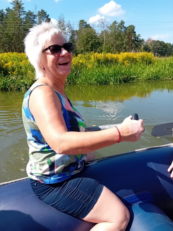 Eine Frau mit kurzen weißen Haaren, bekleidet mit Sonnenbrille, Top und kurzer Hose sitzt lachend auf dem Rand eines blauen Schlauchboots. Sie hält ein Ruder in der Hand. Im Hintergrund ist am Ufer ein Nadelwald erkennbar.