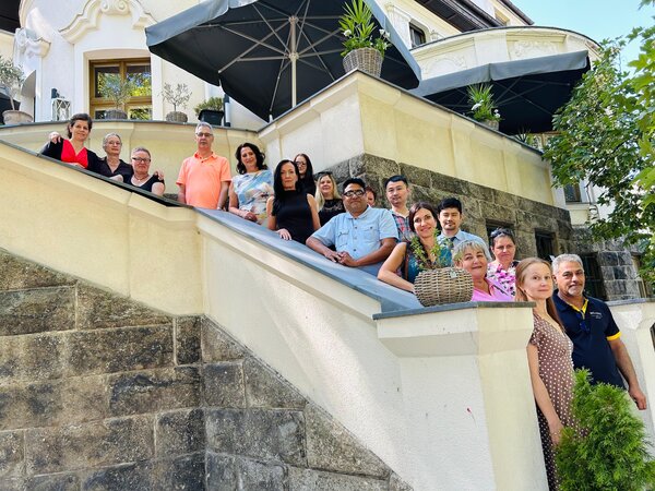 Gruppenfoto von Erwachsenen auf einer Außentreppe vor einem historischen Gebäude mit Terrasse und großen Sonnenschirmen.