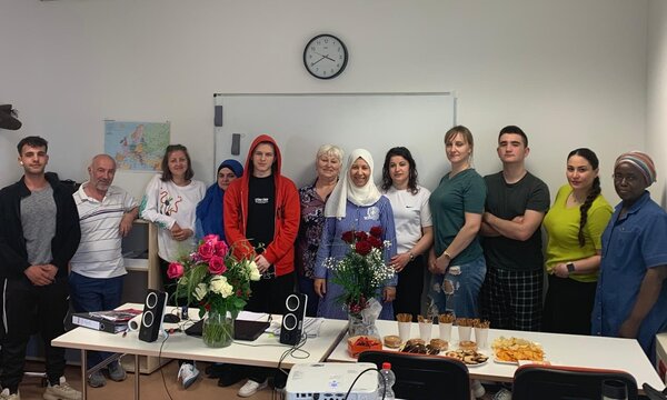 Eine Gruppe von Teilnehmer*innen und Lehrkräften des Integrationskurses steht lächelnd in einem Klassenraum zusammen. Auf dem Tisch vor ihnen befinden sich Blumensträuße, Lautsprecher und ein Buffet mit Snacks und Gebäck. Im Hintergrund eine Weltkarte und eine Wanduhr.