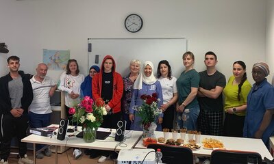 Eine Gruppe von Teilnehmer*innen und Lehrkräften des Integrationskurses steht lächelnd in einem Klassenraum zusammen. Auf dem Tisch vor ihnen befinden sich Blumensträuße, Lautsprecher und ein Buffet mit Snacks und Gebäck. Im Hintergrund eine Weltkarte und eine Wanduhr.