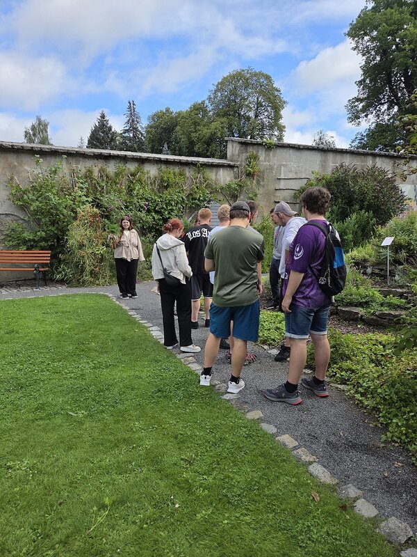 Eine Gruppe von Personen steht auf einem Weg in einem Park. Sie sind von üppigem Grün, Blumenbeeten und einer alten, bewachsenen Steinmauer umgeben. Einige Personen tragen Rucksäcke und scheinen einer Führung zuzuhören.