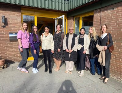 Vor einem Gebäude mit brauner Klinkerfassade und gelbem Eingang stehen sieben Personen nebeneinander und posieren für ein Gruppenfoto. Die Teilnehmenden tragen unterschiedliche, überwiegend helle und dunkle Freizeitkleidung. Im Hintergrund sind große Fenster, ein geöffneter Eingang und ein Briefkasten zu sehen.