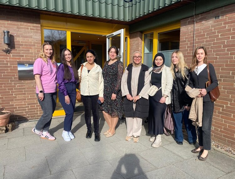 Vor einem Gebäude mit brauner Klinkerfassade und gelbem Eingang stehen sieben Personen nebeneinander und posieren für ein Gruppenfoto. Die Teilnehmenden tragen unterschiedliche, überwiegend helle und dunkle Freizeitkleidung. Im Hintergrund sind große Fenster, ein geöffneter Eingang und ein Briefkasten zu sehen.