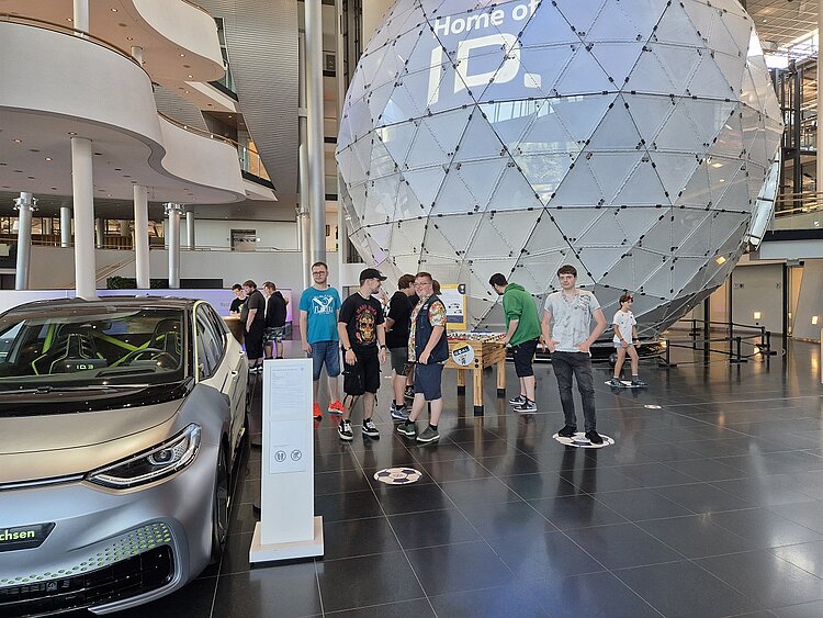 Zu sehen ist links ein silbergraues Auto der Marke VW. Rechts daneben steht eine Gruppe junger Leute. Dahinter befindet sich eine große Glaskugel, die bis zu Decke reicht.