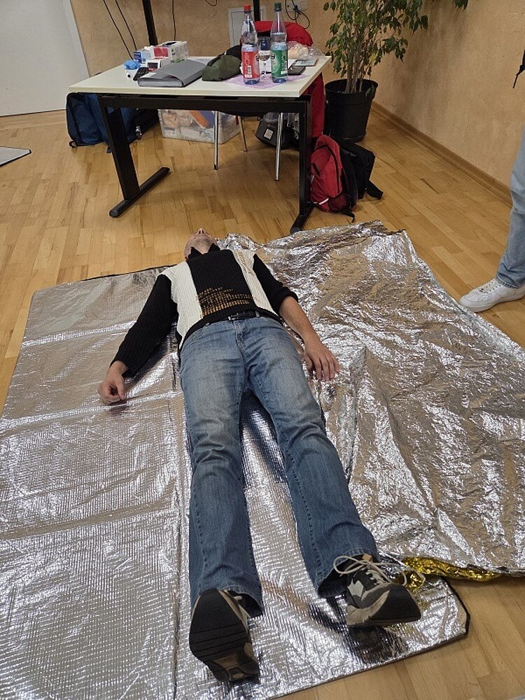 Ein Mitarbeiter der Euro-Schulen liegt auf einer silbernen Rettungsdecke auf dem Boden, während Erste-Hilfe-Materialien auf einem Tisch im Hintergrund bereitstehen. Übungsszene aus einer Erste-Hilfe-Fortbildung in einem Schulungsraum.