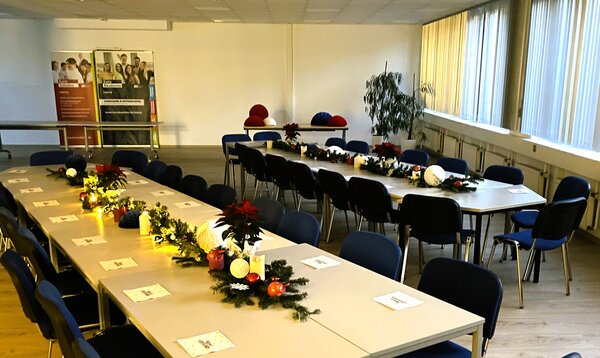 Großes Auditorium mit langen, zu U-Form gestellten Tischen, blauen Stühlen und weihnachtlicher Tischdekoration aus Lichterketten, Tannengrün und roten Sternen.