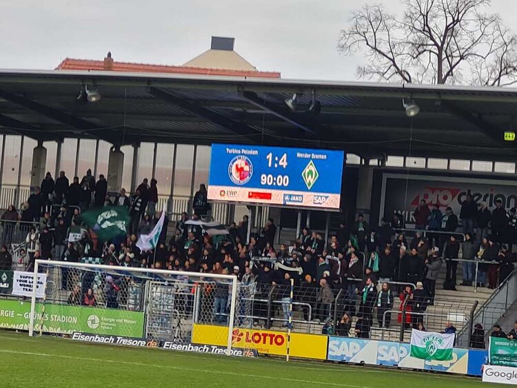 Auf der Tribüne eines mittelgroßen Fußballstadions stehen Menschen  mit Fahnen und Fanschals. Über ihnen thront die Anzeigetafel, die anzeigt, dass das Spiel zwischen dem Gastgeber Turbine Potsdam und Werder Bremen in der 90. Minute 1:4 steht.