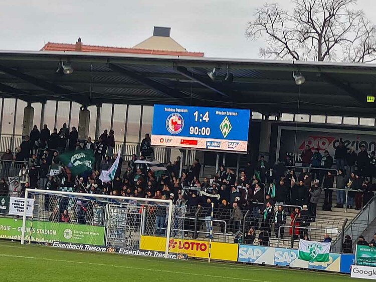 Stadionerlebnis der Extraklasse bei Turbine Potsdam Auf der Tribüne eines mittelgroßen Fußballstadions stehen Menschen mit Fahnen und Fanschals. Über ihnen thront die Anzeigetafel, die anzeigt, dass das Spiel zwischen dem Gastgeber Turbine Potsdam und Werder Bremen in der 90. Minute 1:4 steht.