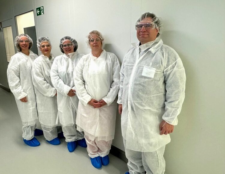 Fünf Teilnehmende stehen nebeneinander an einer hellen Wand in einem Innenraum. Sie tragen weiße Schutzanzüge, Haarnetze und blaue Überschuhe. Die Gesichter sind unkenntlich gemacht. An der Wand befindet sich ein grünes Schild mit einem weißen Symbol.