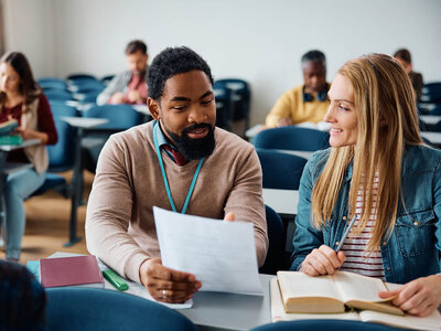 Ein Mann und eine Frau sitzen an einem Tisch in einem Klassenzimmer oder einer Lernumgebung. Der Mann, der ein Namensschild um den Hals trägt, hält ein Blatt Papier und scheint es der Frau zu zeigen oder zu erklären. Die Frau, die ein gestreiftes Hemd und eine Jeansjacke trägt, hält ein offenes Buch und einen Stift und scheint zuzuhören oder Notizen zu machen. Im Hintergrund sind andere Personen zu sehen, die ebenfalls an Tischen sitzen und arbeiten oder lernen.