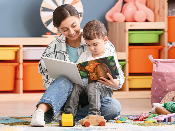 Situation in einer Kita: Eine Erzieherin und ein Kind sitzen auf dem Boden in einem farbenfrohen Raum und lesen gemeinsam ein Bilderbuch. Im Hintergrund sind bunte Spielzeugkisten, ein Regal mit Spielzeug und ein Stoffbehälter zu sehen. Auf dem Boden liegen verschiedene Spielsachen verstreut, darunter ein Holzauto und ein gelber Spielzeuglaster.