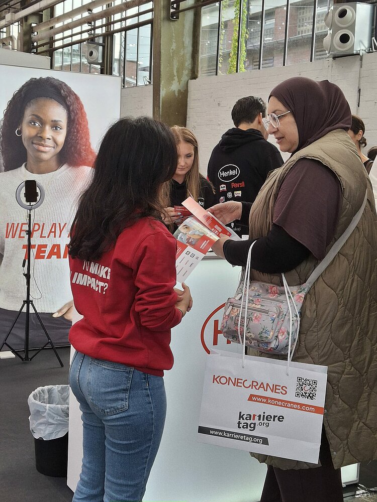 Eine Teilnehmerin informiert sich an einem Stand und wird dort beraten. Über den Arm hängt eine Papiertüte mit Messelogo.