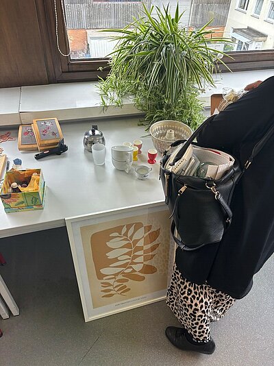 Unser Nachhaltigkeitsbasar ist immer ein Highlight Eine Frau steht an einem Tische, der mit verschiedenen Utensilien wie beispielsweise Kaffeegeschirr bestückt ist, und betrachtet die Gegenstände. Auch ein großformatiges Bild lehnt an dem Tisch. Auf der Fensterbank steckt eine große Grünlilie, das Fenster dahinter gibt den Blick in einen Hinterhof frei.