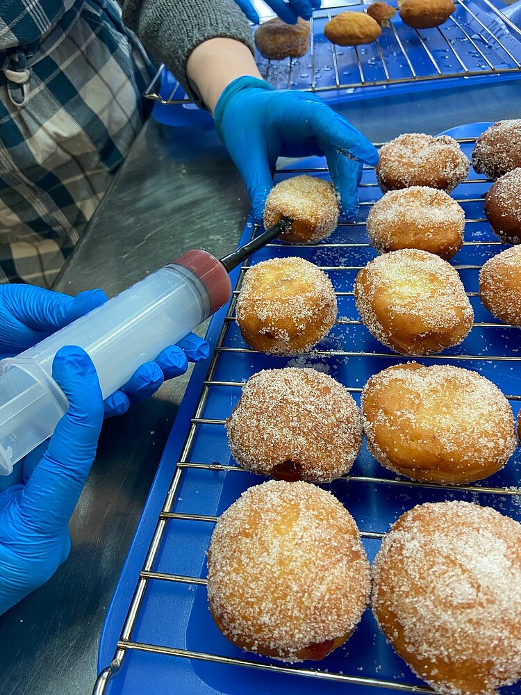 Kein Karneval in Bielefeld Eine Person mit blauen Einweghandschuhen füllt mit einer Spritze Marmelade oder eine andere Füllung in frisch gebackene Krapfen. Die Krapfen liegen auf einem blauen Gitter zum Abkühlen.