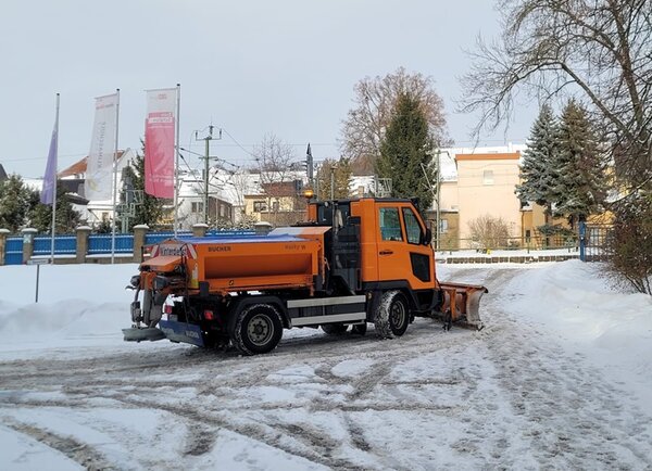 Ein orangefarbener Schneeräumfahrzeug des Bauhofs Hohenstein-Ernstthal steht auf einem verschneiten Hof. Das Fahrzeug ist mit einem großen Schneepflug an der Front und einem Streuaufsatz auf der Ladefläche ausgestattet. Im Hintergrund sind Fahnenmasten mit Bannern der Euro-Schulen Hohenstein-Ernstthal sowie Wohnhäuser und Bäume zu sehen. Der Boden ist teilweise geräumt und mit Schneeresten bedeckt.