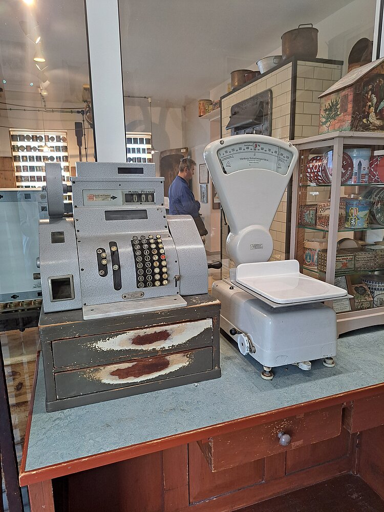 Innenansicht eines Museums mit einer alten Registrierkasse und einer mechanischen Waage auf einem Tisch. Im Hintergrund sind Personen und historische Gegenstände zu sehen.