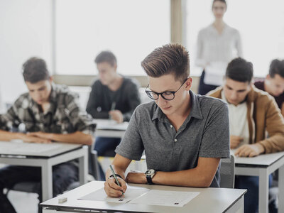 Ein junger Mann mit Brille und einem grauen Hemd sitzt an einem Schreibtisch in einem Klassenzimmer und schreibt auf ein Blatt Papier. Im Hintergrund sind weitere Schüler zu sehen, die ebenfalls an ihren Tischen sitzen und arbeiten. Eine Lehrerin steht im Hintergrund und beobachtet die Klasse. Der Raum ist hell und hat große Fenster, die Tageslicht hereinlassen.