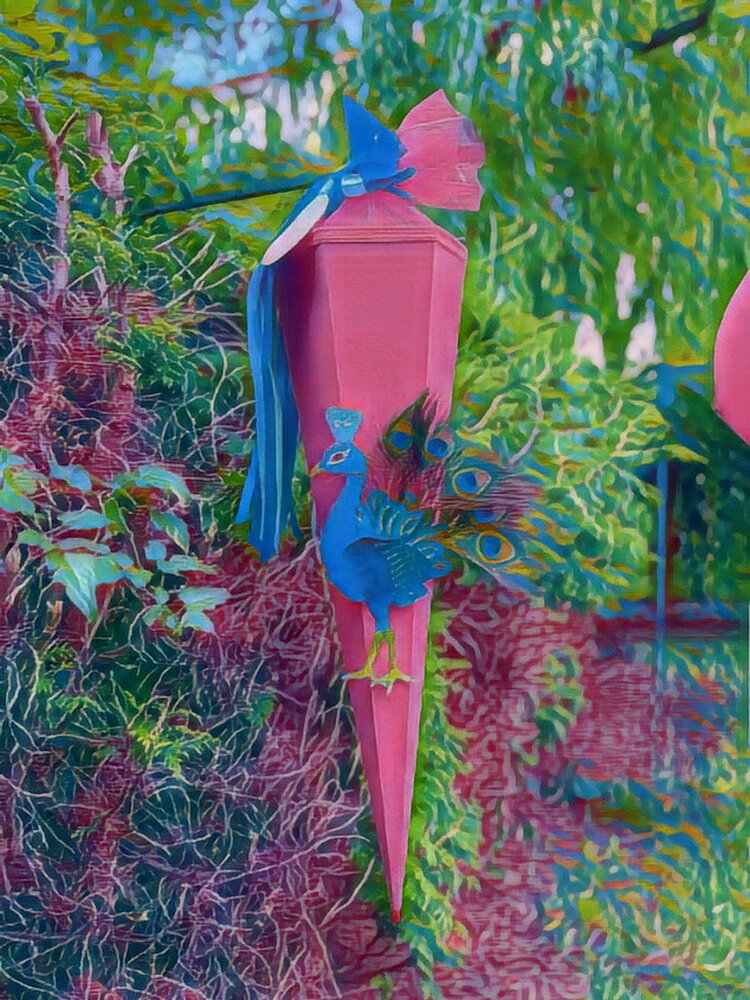 Eine pinke Schultüte mit einem dekorativen Pfauenmotiv und blauer Schleife, die in einen üppig bunten Busch aufgehängt ist, dessen Blätter den Farben der Schultüte entspricht.